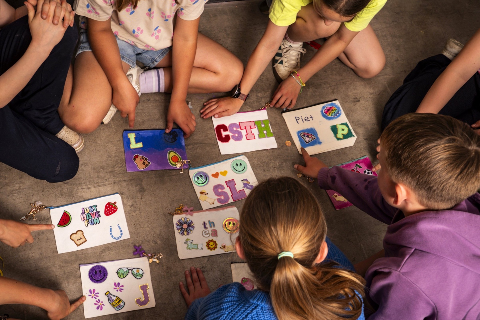 Kinder sitzen im Kreis auf dem Boden und präsentieren stolz ihre selbst verzierten Stoffmäppchen mit bunten Patches im Kreativraum der Werkstadt Vechta